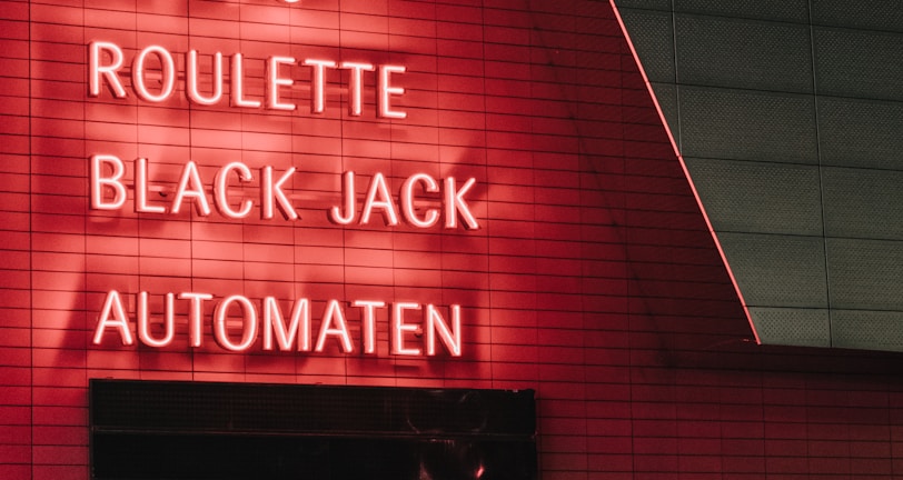 Red neon letters are mounted on a dark tiled wall, displaying the words Poker, Sic Bo, Roulette, Black Jack, and Automaten.