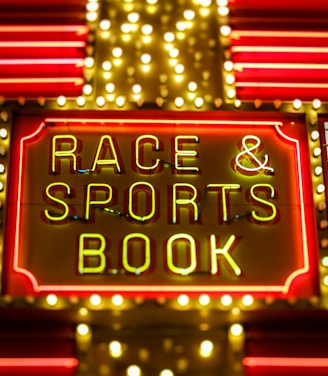 Bright neon lights on a marquee sign display the words 'RACE & SPORTS BOOK'. The sign is surrounded by rows of yellow and white lights against a red background, evoking a vibrant, energetic atmosphere typically associated with casinos or entertainment venues.