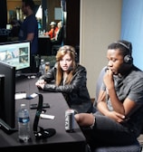 Two people are seated at a table with gaming monitors, wearing headsets. They appear to be participating in an esports broadcast or commentary. The environment is dimly lit with additional equipment and people visible in the background. A large blue banner with the word 'Frostbite' and a stylized graphic of a wolf is displayed.