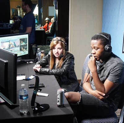 Two people are seated at a table with gaming monitors, wearing headsets. They appear to be participating in an esports broadcast or commentary. The environment is dimly lit with additional equipment and people visible in the background. A large blue banner with the word 'Frostbite' and a stylized graphic of a wolf is displayed.