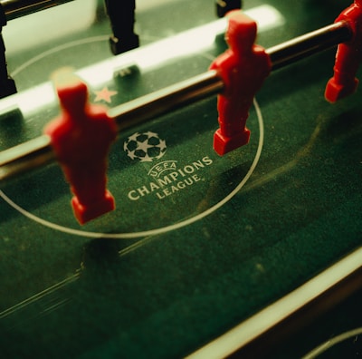A foosball table is shown with red and black player figures positioned on the rods. The surface features the UEFA Champions League logo, indicating a themed table design.