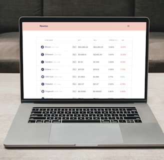 A laptop on a table displaying a cryptocurrency trading platform. The screen lists various cryptocurrencies along with their buy and sell values, spread percentages, and 24-hour change percentages. The keyboard and touchpad of the laptop are visible, set against a neutral background with a sofa in the back.