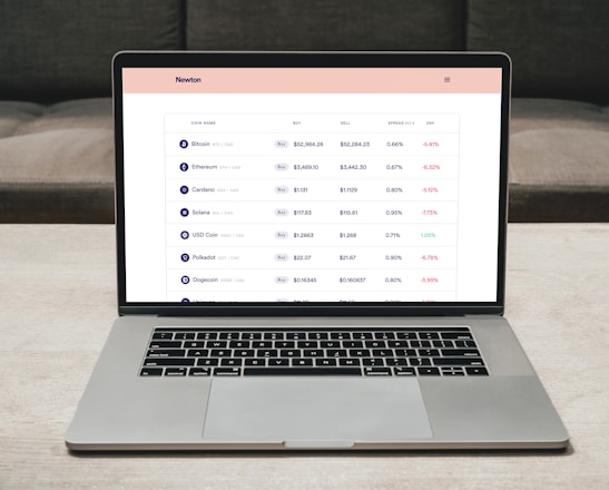 A laptop on a table displaying a cryptocurrency trading platform. The screen lists various cryptocurrencies along with their buy and sell values, spread percentages, and 24-hour change percentages. The keyboard and touchpad of the laptop are visible, set against a neutral background with a sofa in the back.