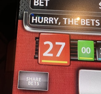 A digital display screen showing a casino betting interface, with text prompting to hurry bets and a highlighted number 27 in red. There are options for betting amounts and a 'share bets' button.