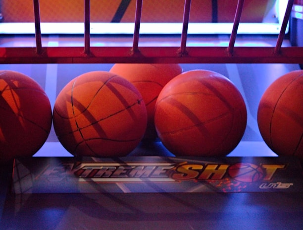 Several basketballs are aligned in a row on a basketball arcade machine platform. The environment has a colorful and illuminated backdrop with a mix of red and blue lighting.